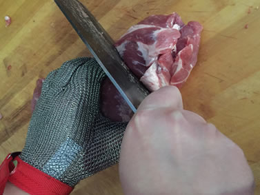 A hand wearing chainmail glove is holding a piece of meat and the other hand is cutting the meat.